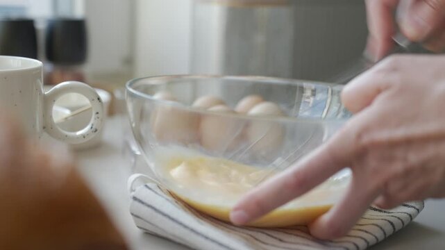Hands whisk eggs in bowl on kitchen counter, with mug and extra eggs nearby. Concept of breakfast preparation and simple cooking