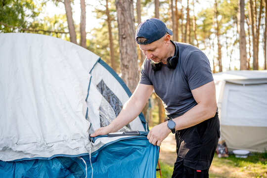 Man builds a blue camping tent in a sunny forest area during a calm morning.
