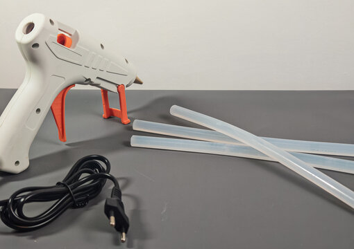 A professional high-resolution commercial product shot featuring a white electric hot melt glue gun with its black power cord and translucent adhesive sticks arranged neatly on a grey surface.