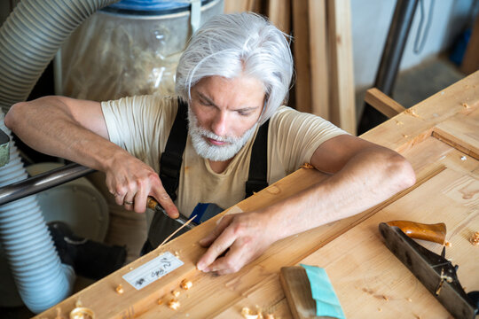 Focused craftsman carpenter professionally working with chisel tool, carving wood layers on door, concentrated handyman woodworker restoring vintage furniture, engaged in creative renovation process.