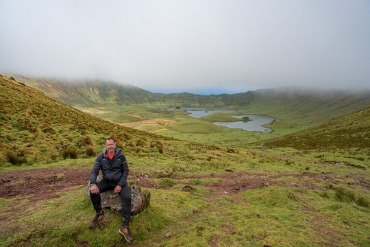 A white European man in a black coat sits on a stone with the bottom of a cauldron. A white European man in a black coat sits on a stone with the bottom of a cauldron on a foggy day, Corvo Island, Azo