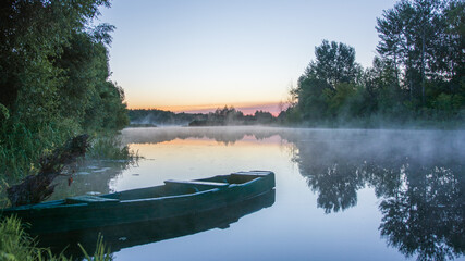 Obraz premium A serene wooden boat floats on a misty, calm river surrounded by lush green trees during a peaceful, glowing sunrise