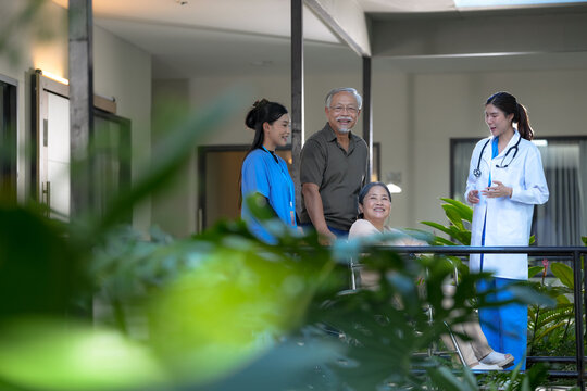 Lifestyle of aging: Senior Asian woman seated in a wheelchair outdoors with her family and a smiling nurse, enjoying a gentle walk through a leafy garden, captured with care