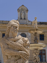 Obraz premium Compelling close-up perspective of marble sculpture at Fontana Pretoria in Palermo, Sicily, against the historic Palazzo Pretorio clock tower and the statue of Santa Rosalia under a clear blue sky.