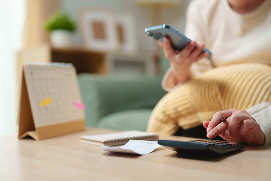 Woman using smartphone and calculator to manage household budget with receipts, notebook and calendar at home. Personal finance, expense tracking, bill payment, budgeting and financial planning