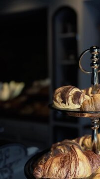 Croissants sit on a metallic stand in a bakery. The soft golden pastries show layers of dough and chocolate filling. The background features shelves filled with more baked goods.