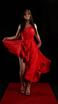 Stunning Black-Haired Woman in Ruby Red Dress and High Heels Gracefully Posing Against a Black Background, Captured in Super Slow Motion. Vertical Aspect Ratio.