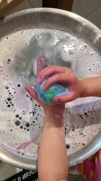 Vertical UGC about cosmetics. Wet hands rubbing turquoise bath bomb inside round metal basin filled with dense white suds and bubbles, with glossy soap film, bright reflections. Indoor. Kids hands