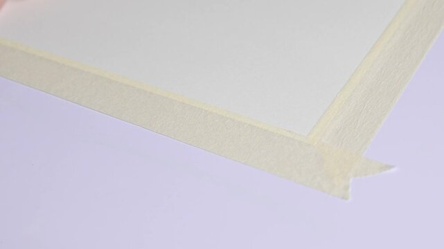 Artist hands taping paper sheet to drawing board with masking tape before painting, preparing art workspace for watercolor illustration, close-up creative process