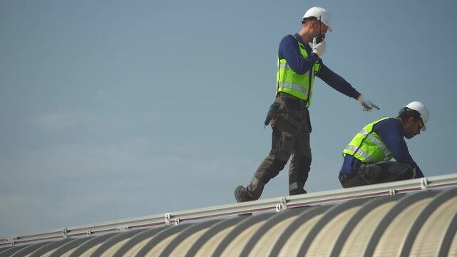 Two workers stand silhouetted on the roof ridge against the bright sky, pausing between tasks during the rooftop solar panel installation, filmed during a large scale rooftop solar power