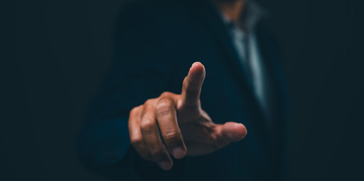 A person in a dark suit points forward with their index finger against a dark background, emphasizing focus and interaction.