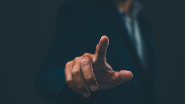 A person in formal attire points forward, with focus on the extended index finger against a dark blurred background.