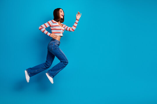 Happy young woman jumps in blue studio wearing striped top and jeans showing chic casual lifestyle