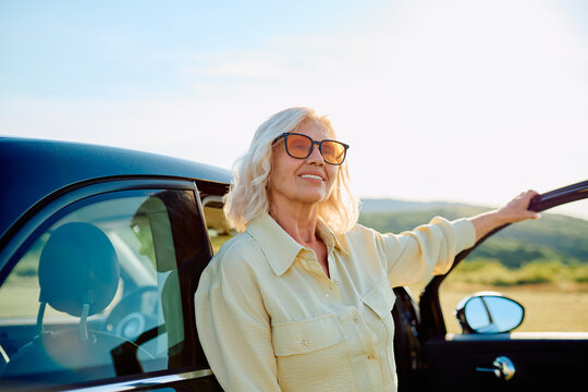 Smiling senior woman wearing sunglasses and leaning on her car, enjoying a sunny day outdoors and discovering new places