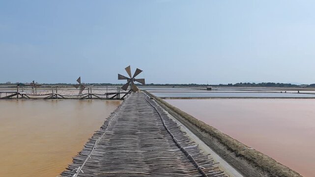 Slow panning right video of a scenic traditional salt farm with a bamboo bridge and wooden windmills under a clear blue sky.