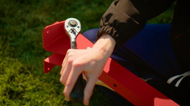 DIY builder adjusting socket wrench on metal frame joint in garden setting. Home improvement installation process with red painted parts and manual labor. Outdoor construction concept with hands