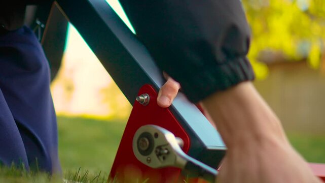 Hands using ratchet wrench on angled metal frame during garden construction. Red bracket and gray beam assembly with bolt connection and manual pressure. DIY installation concept with technician