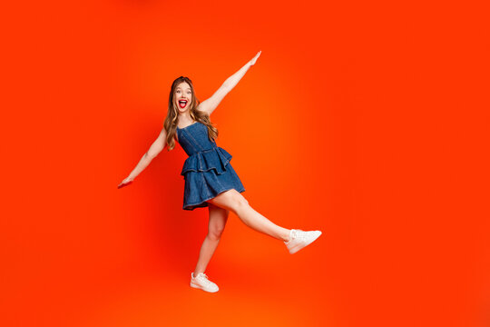 Joyful young woman in denim dress posing energetically on vibrant orange background with white sneakers and bright red lipstick