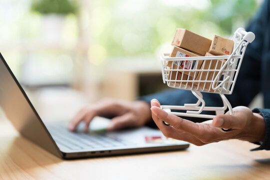 Shopping online concept. Close up of hands holding shopping cart model with many boxes on table during using laptop computer on table