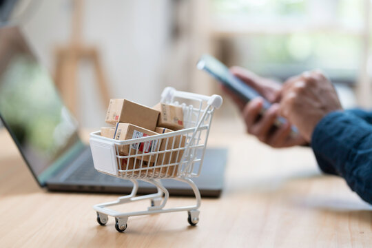 Shopping online concept. Close up of hands using mobile phone, laptop computer and shopping cart model with many boxes on table