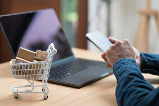 Shopping online concept. Close up of hands using mobile phone, laptop computer and shopping cart model with many boxes on table