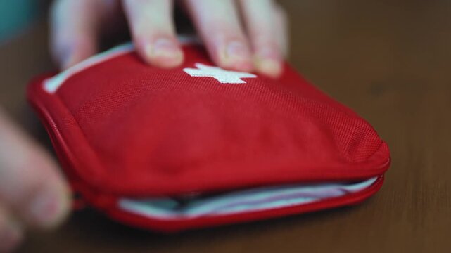 Close up of hands packing and closing a red portable first aid kit pouch full of medical pills on a wooden table.