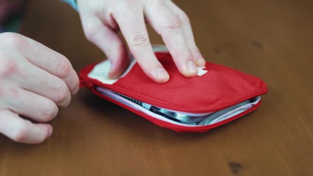 Close up of hands packing and closing a red portable first aid kit pouch full of medical pills on a wooden table.
