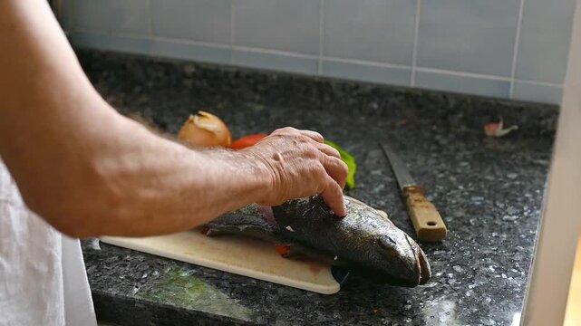 Male hands preparing fresh whole meagre fish on wooden cutting board, making cuts before stuffing with vegetables for healthy Mediterranean seafood recipe and homemade oven baked dinner