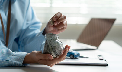 Fototapeta premium Businesswoman hand putting coin in glass jar with dollar bills for saving to buy a new car. Auto loan, vehicle insurance, and financial planning investment concept at office desk.