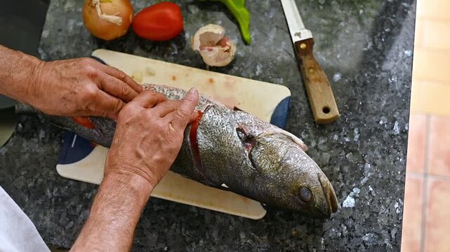 Top view of male hands stuffing fresh tomato slices into cuts of whole meagre fish, preparing healthy Mediterranean seafood recipe with fresh ingredients for oven baking