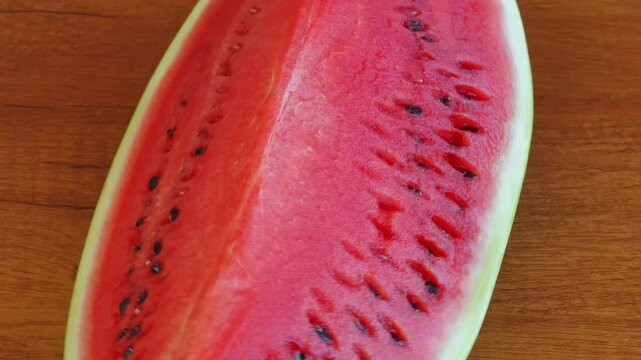 A slice of ripe red watermelon rotating on a textured wooden table surface with a static camera view. High speed fps footage for summer food commercial, organic fruit and refreshing dessert video. 