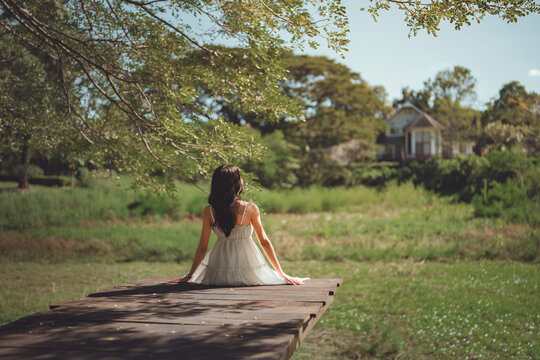 Back view of happy woman in white dress sitting on wooden pier at green park, beautiful female traveler relaxing and looking at view during summer vacation for travel tourism, mental wellness