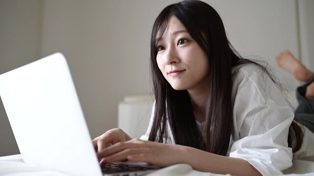 ベッドでノートパソコンを操作して微笑む20代日本人女性