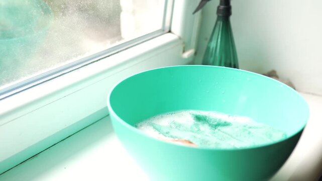 Close up of a blue basin with soapy water and spray bottle on a window sill