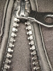 Detailed close-up of a metal zipper, partially unzipped, showcasing its teeth and pull on a textured dark grey fabric © Rayka
