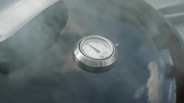 An analog thermometer gauge on a closed barbecue grill lid measures internal heat. Smoke drifts over the black surface during backyard cookout. Temperature control ensures precise smoking.