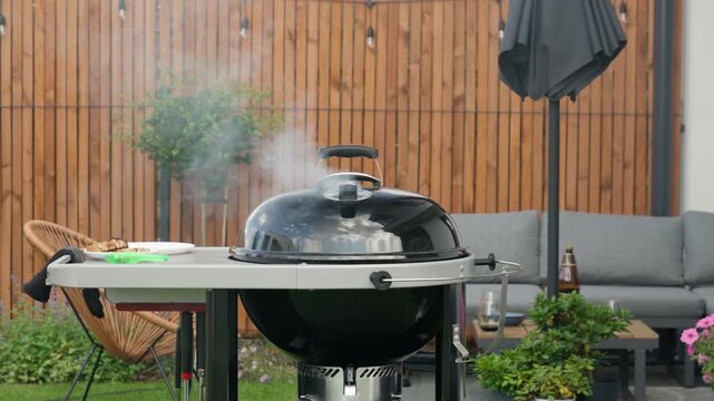 Thick smoke rises from a closed charcoal grill on an outdoor terrace during summer barbecue. Food slowly cooks inside kettle grill. Modern backyard patio setup for family outdoor dining.