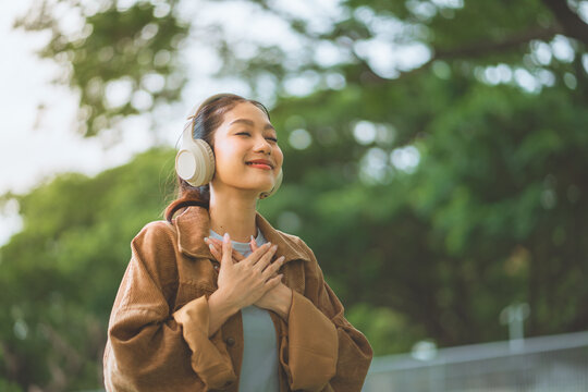 Happy Asian woman wearing headphones with eyes closed and hands on chest at green park, smiling female listening to music, audio podcast, or breathing fresh air for mental health, mindfulness wellness