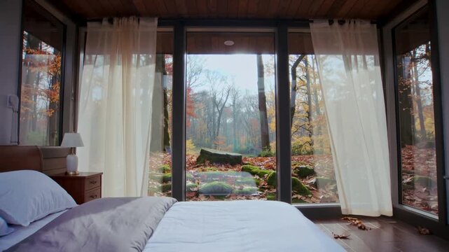 Camera pulling back revealing bedroom with full-height glass wall in forest cabin, showing view