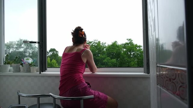 A woman looks out the window at the rain while sitting in the room.
