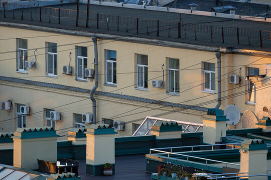 White air conditioners mounted under windows of pale yellow building. Metal drain pipes run vertically down wall. Dark green roof parapet with decorative trim below. Urban architectural detail.