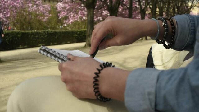 An artist draws a botanical illustration in a spiral notebook while sitting outdoors. The frame focuses on the person's hands holding a pen and sketching against a park background.