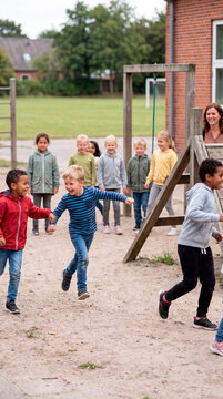 Joyful children racing across sandy schoolyard as teacher watches during recess