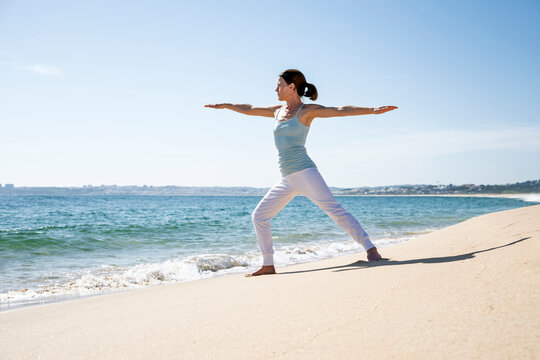 woman practices yoga in Warrior II pose on a sunny sandy beach