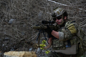 Fototapeta premium Tactical operator in camouflage gear rests with a rifle on rocky terrain during outdoor field training. Concept of military exercise, security, readiness, and tactical operations.