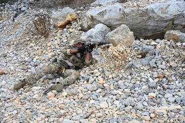 Obraz premium Focused military infantry soldier lying prone on rugged rocky terrain aiming a tactical assault rifle during a dangerous combat training mission for national defense and special security.