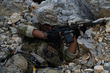 Obraz premium Focused military infantry soldier lying prone on rugged rocky terrain aiming a tactical assault rifle during a dangerous combat training mission for national defense and special security.