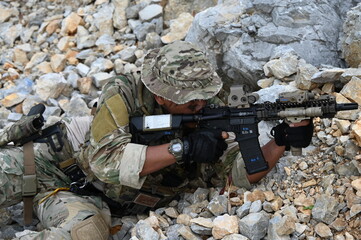 Obraz premium Focused military infantry soldier lying prone on rugged rocky terrain aiming a tactical assault rifle during a dangerous combat training mission for national defense and special security.