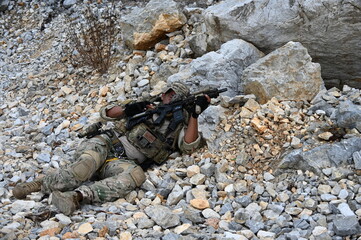 Obraz premium Focused military infantry soldier lying prone on rugged rocky terrain aiming a tactical assault rifle during a dangerous combat training mission for national defense and special security.