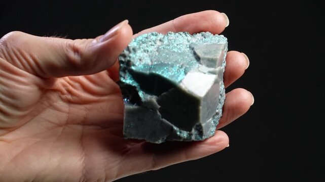 Presenting hand rotating and tilting mineral specimen at black backdrop, revealing turquoise crust